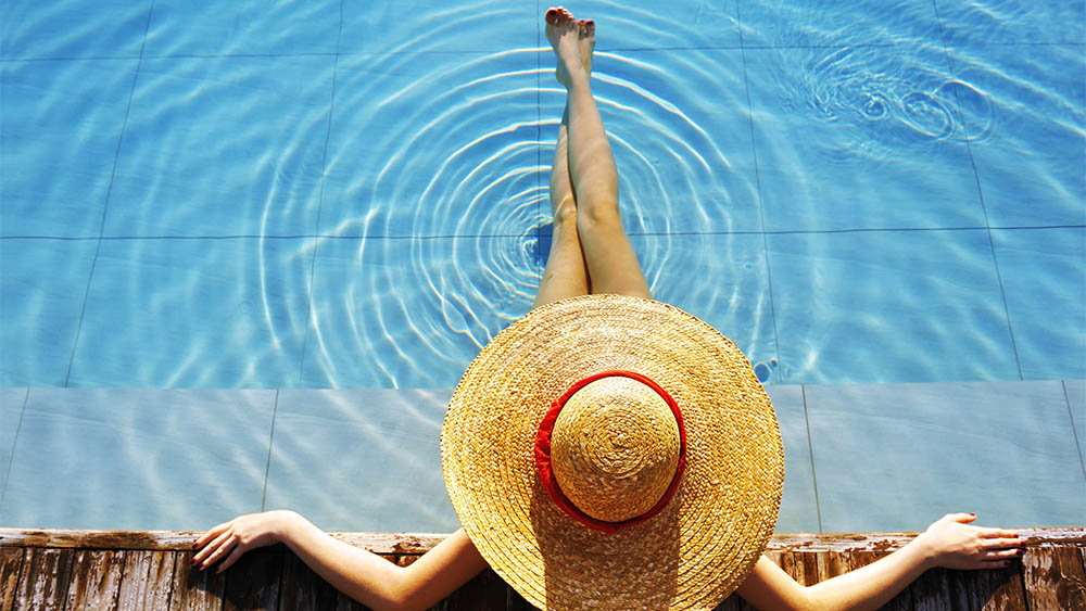 Vista de cima de uma mulher com chapéu sentada na borda de uma piscina.