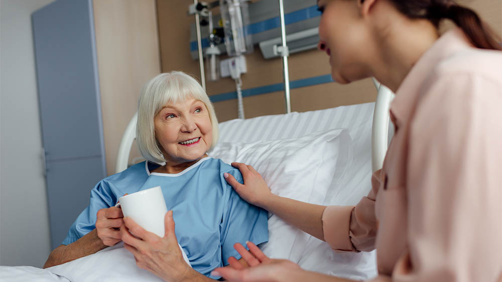 Mulher mais velha sorrindo sentada em cama de hospital com mulher conversando ao lado.